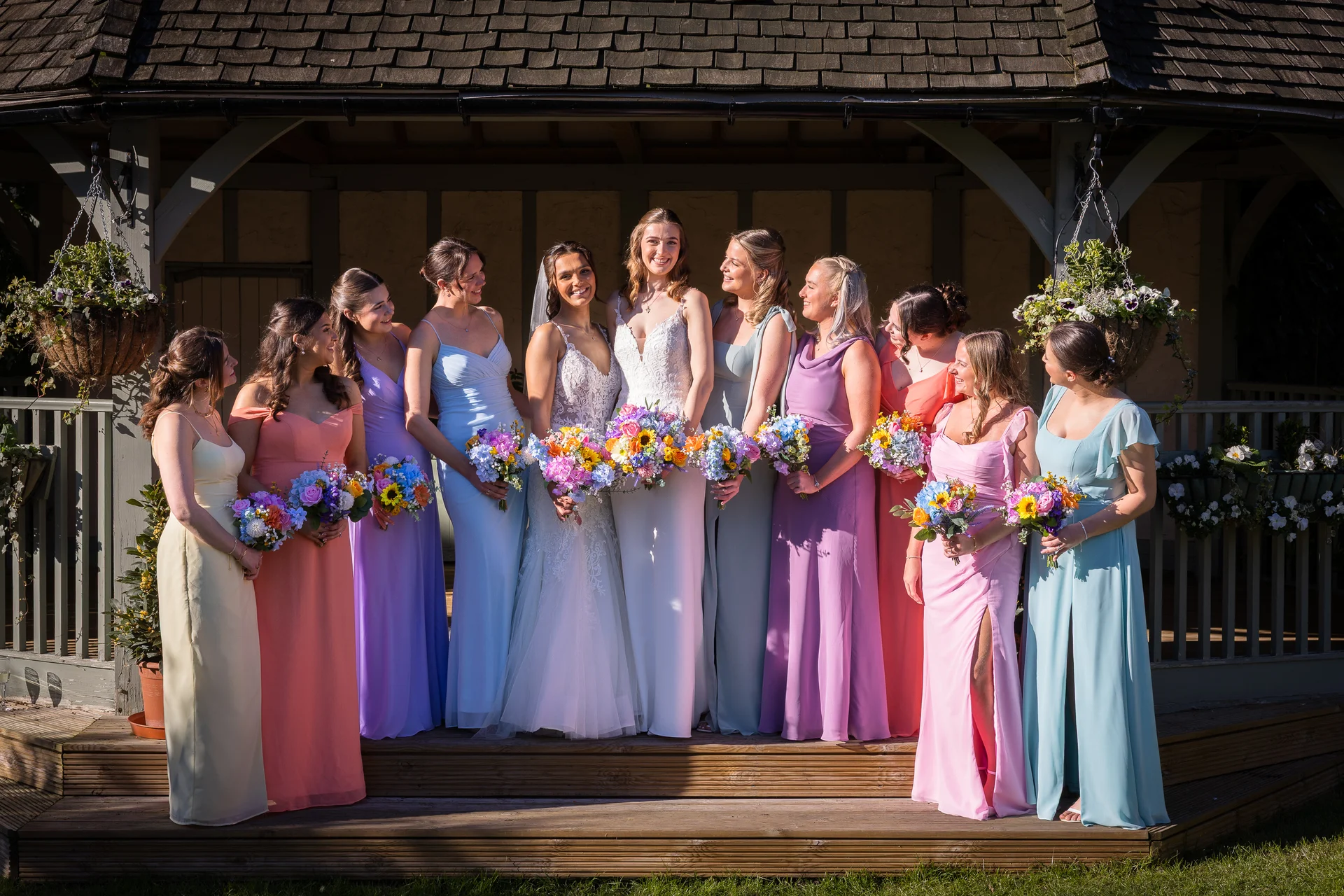 The two smiling brides pose happily with their large bridal party of ten bridesmaids in mismatched pastel-coloured dresses on wooden steps outside a gazebo.