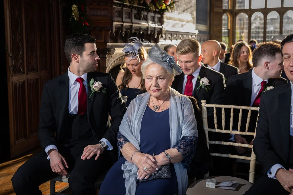 An elderly female guest wearing a navy dress and grey shawl sits attentively among seated guests and groomsmen during a formal wedding ceremony inside a wood-panelled hall.