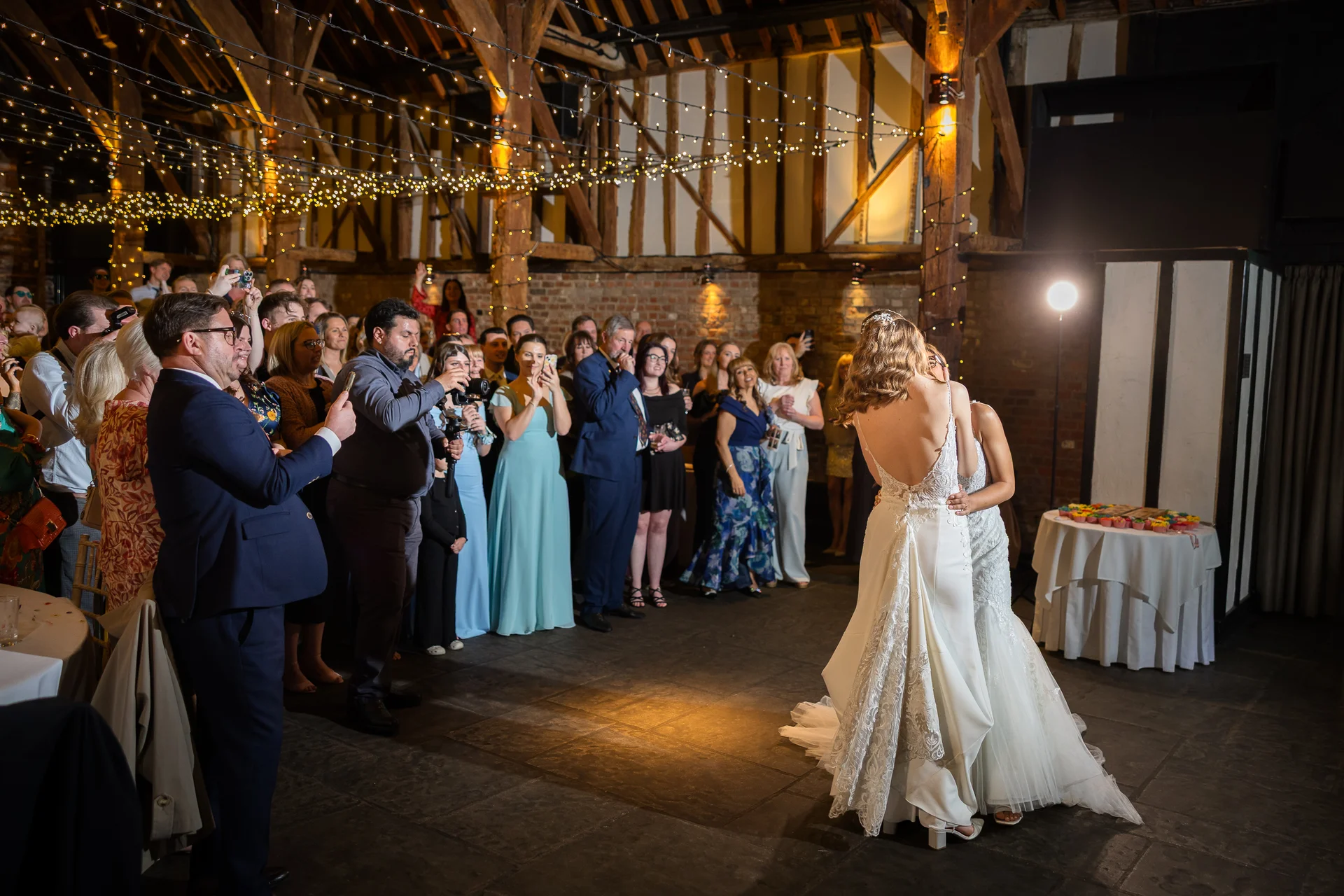 Two brides share a tender first dance in an oak framed barn illuminated by a canopy of fairy lights while wedding guests look on and capture the moment.