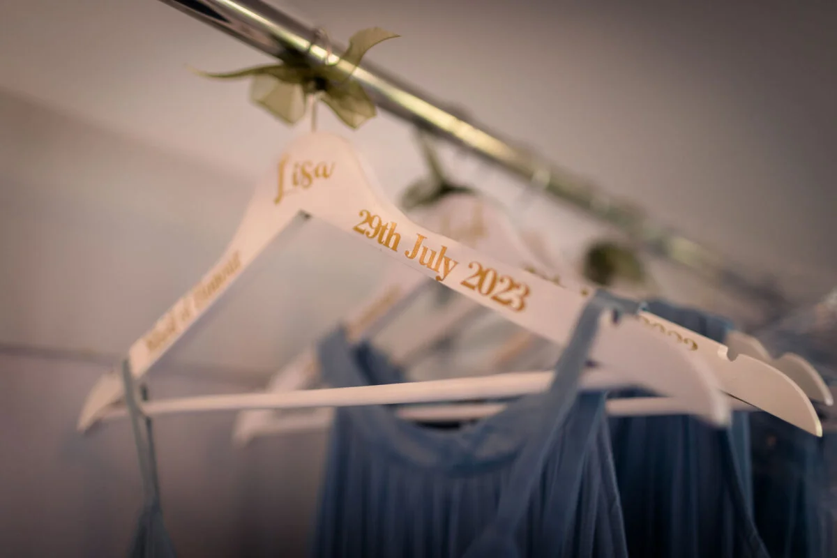 A personalised white wooden hanger with gold lettering featuring a name and wedding date hangs with dusty blue bridesmaid dresses.