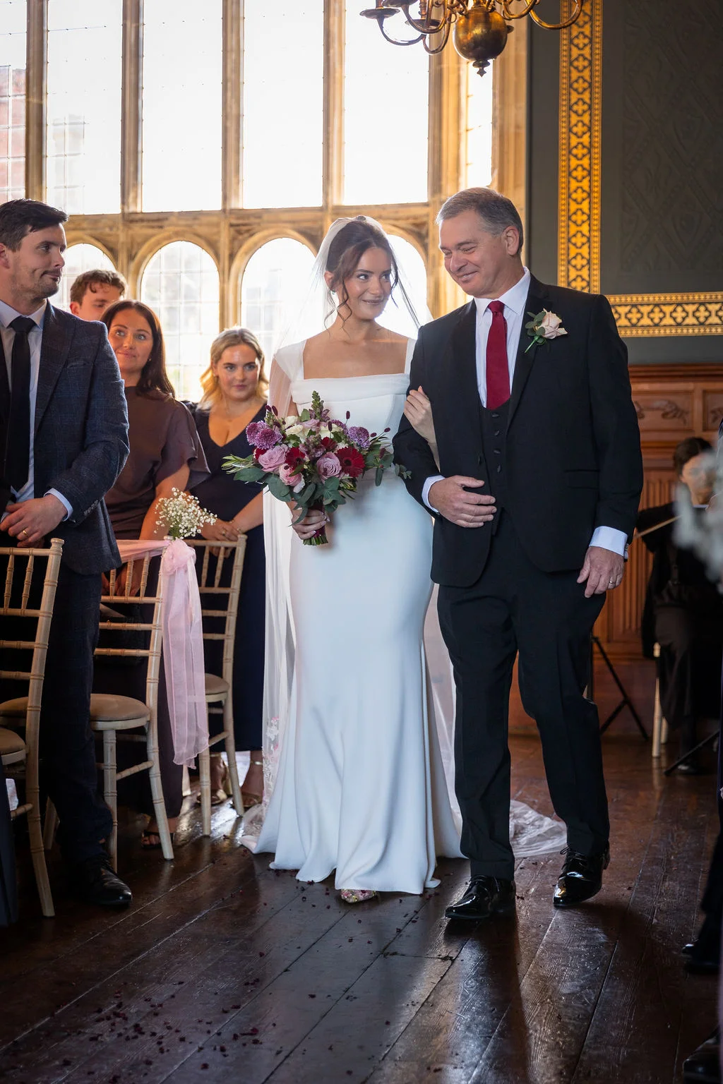 The bride walks down the aisle arm in arm with her father inside a grand, wood-panelled hall with large windows.