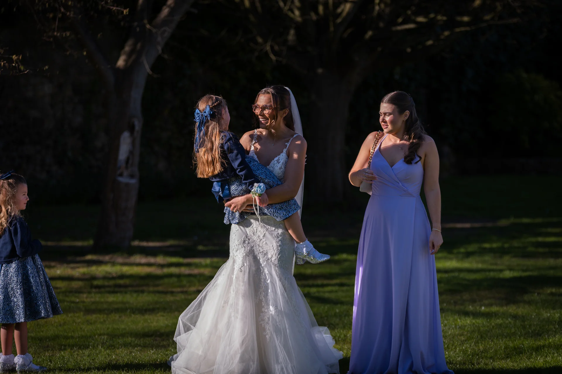 A smiling bride wearing glasses and a lace wedding dress holds a young flower girl in an outdoor garden setting, while a bridesmaid in a lilac dress looks on.