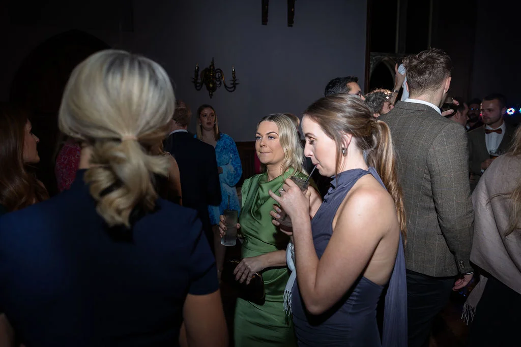 Wedding guests socialise and enjoy drinks during the evening reception inside a dimly lit venue captured with direct flash.
