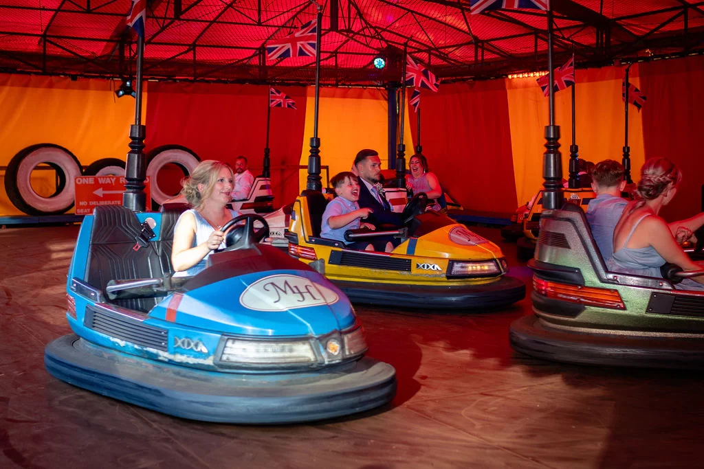 Wedding guests laugh and smile while driving bumper cars inside a brightly lit vintage fairground attraction decorated with union jack flags.