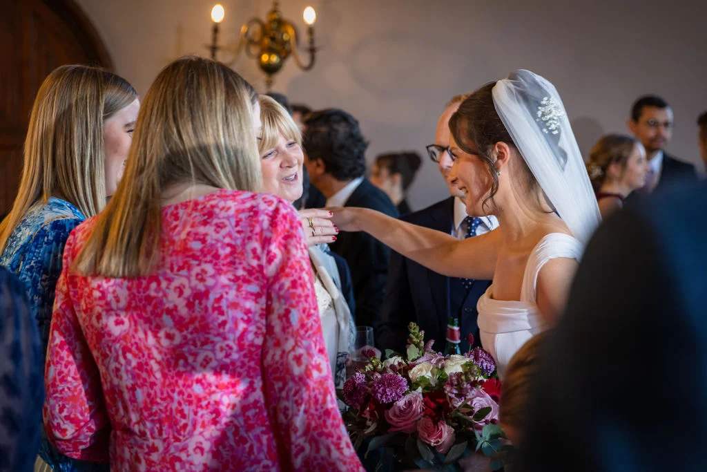 A smiling bride wearing a white gown and veil holds a vibrant floral bouquet while warmly greeting guests during the wedding reception.