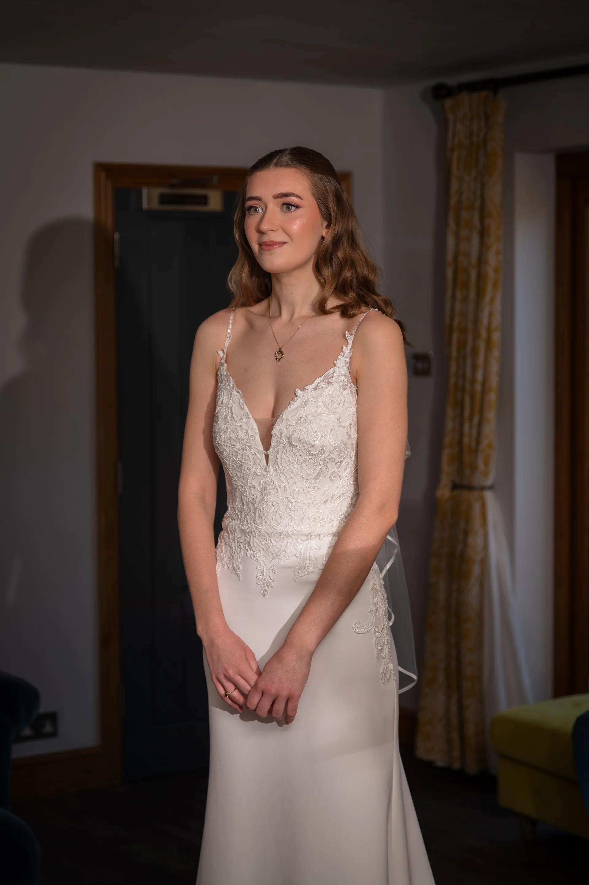 A stunning bride poses indoors wearing a delicate lace and crepe gown with a plunging V-neckline and spaghetti straps, featuring soft half-up wavy hair and a subtle pendant necklace.