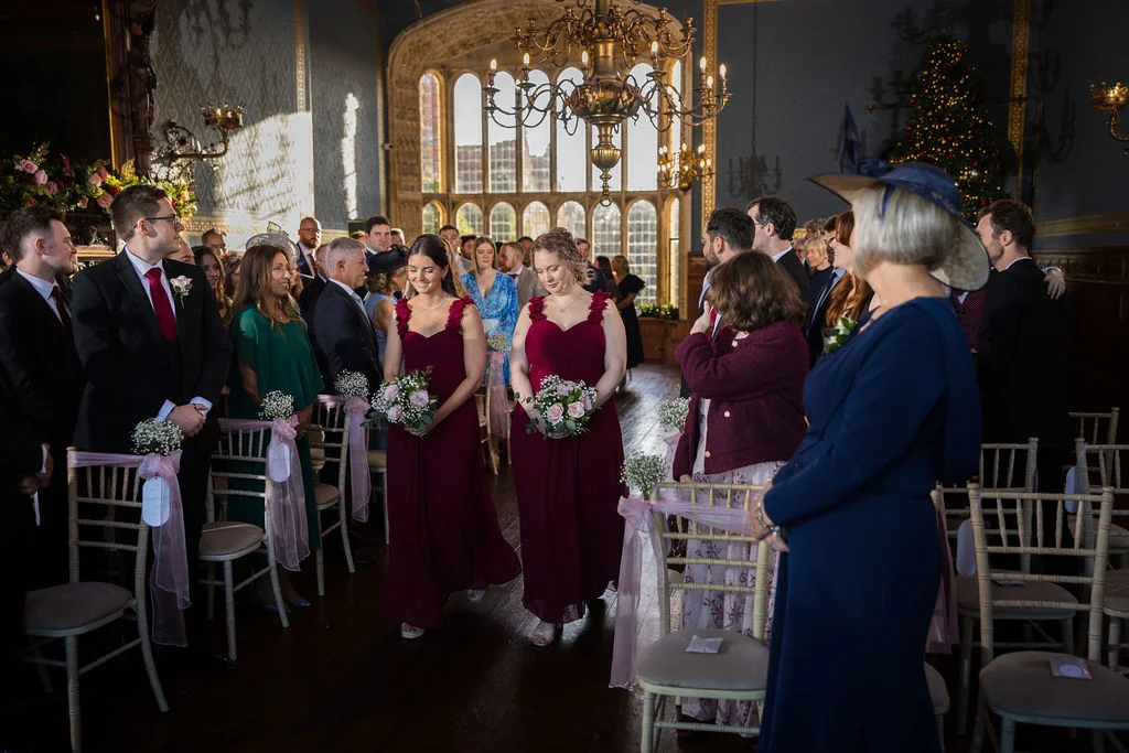Two bridesmaids in deep burgundy dresses walk down the aisle carrying posy bouquets during a wedding ceremony inside a grand stately hall with large arched windows.