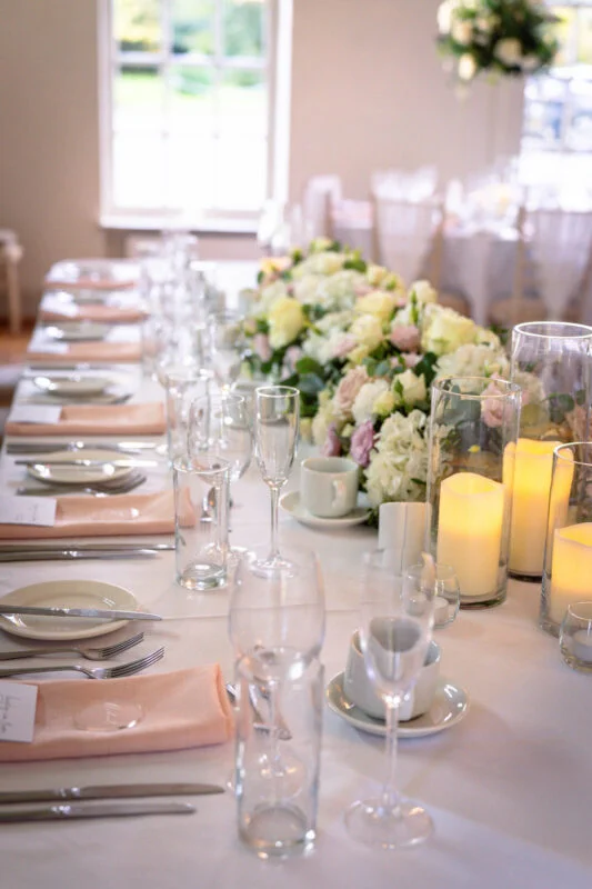 An elegant wedding reception tablescape features crisp white linens, peach napkins, glowing pillar candles in glass cylinders, and a lush floral runner of white and blush blooms.