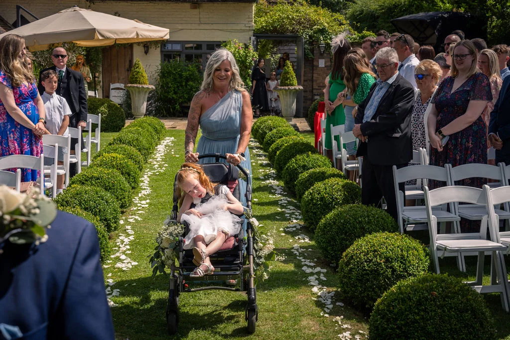 A smiling woman wheels a young flower girl in a decorated pushchair down an outdoor ceremony aisle lined with topiary bushes and scattered white petals.