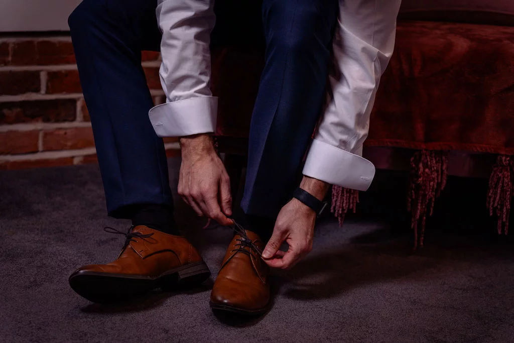 A groom in a white shirt and dark trousers sits down to tie his tan leather brogues while getting ready on the morning of the wedding.