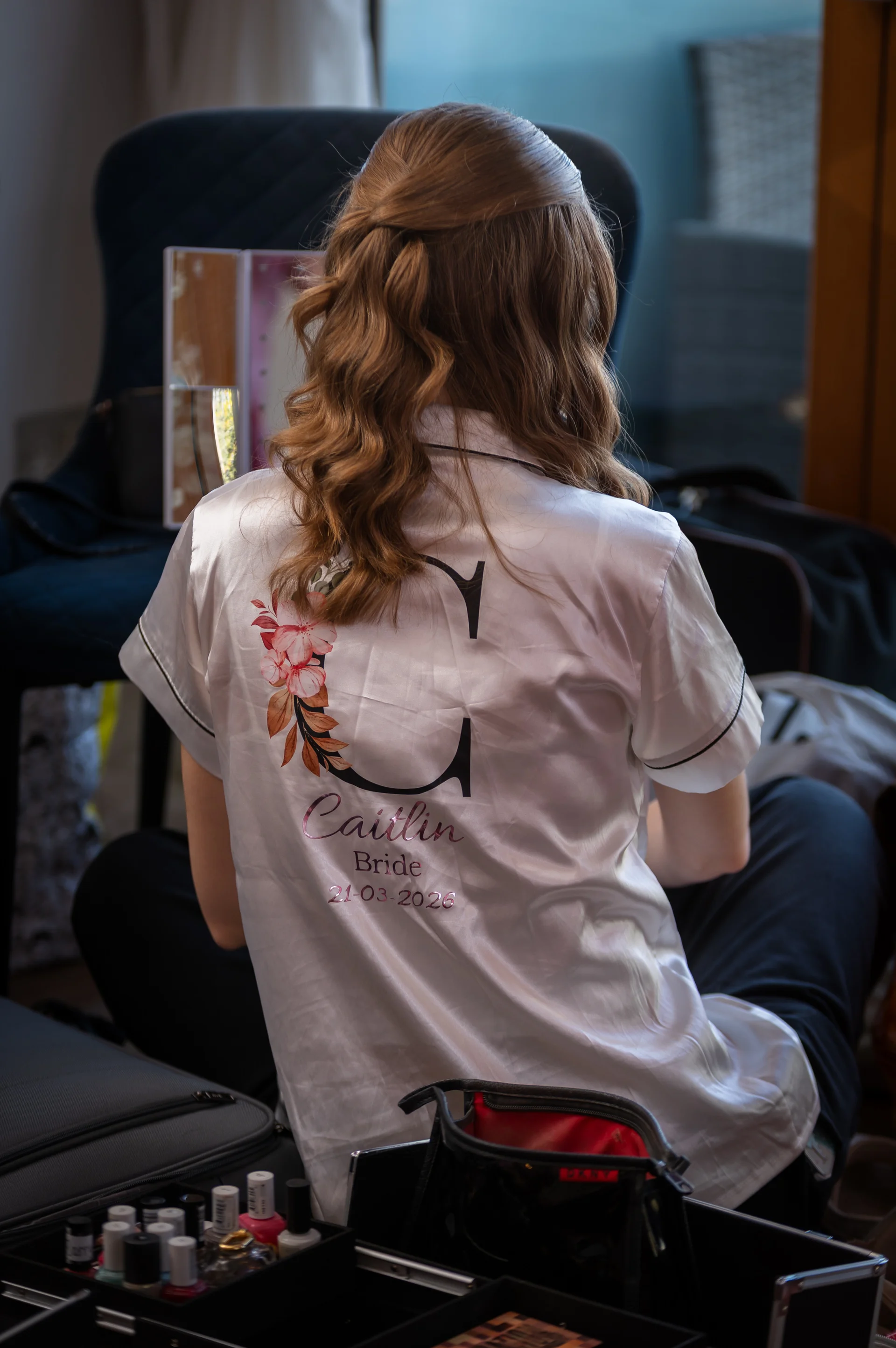 A bride sits with her back to the camera, wearing a personalised white satin top with soft waves in her hair during wedding morning preparations.