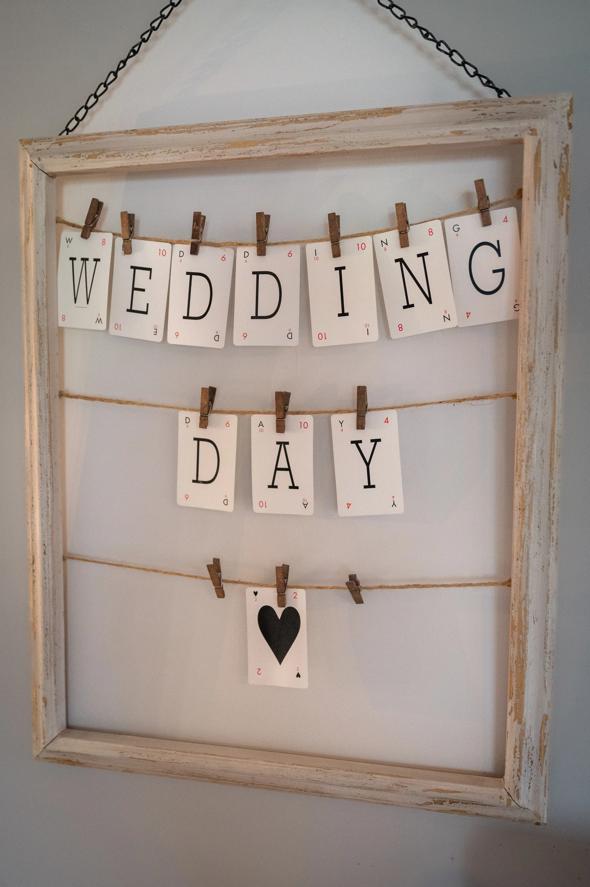 A rustic distressed wooden frame hangs on a wall displaying playing cards clipped to string spelling out the words wedding day.