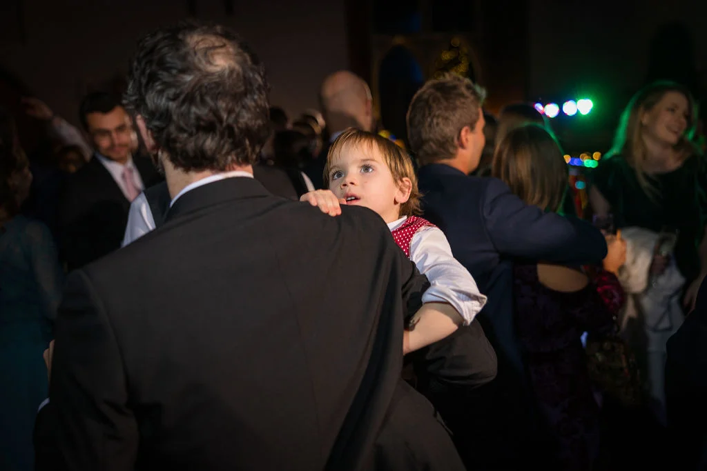 A young page boy looks up curiously while being held by a man on a crowded wedding dance floor.