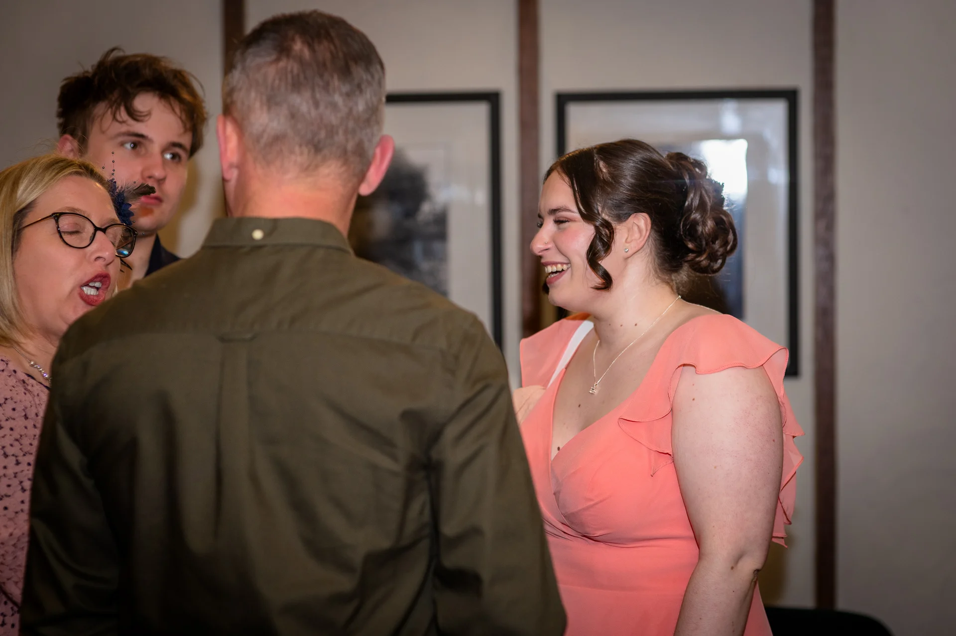 Wedding guests mingling and laughing together indoors, with a woman in a peach v-neck dress smiling during conversation.