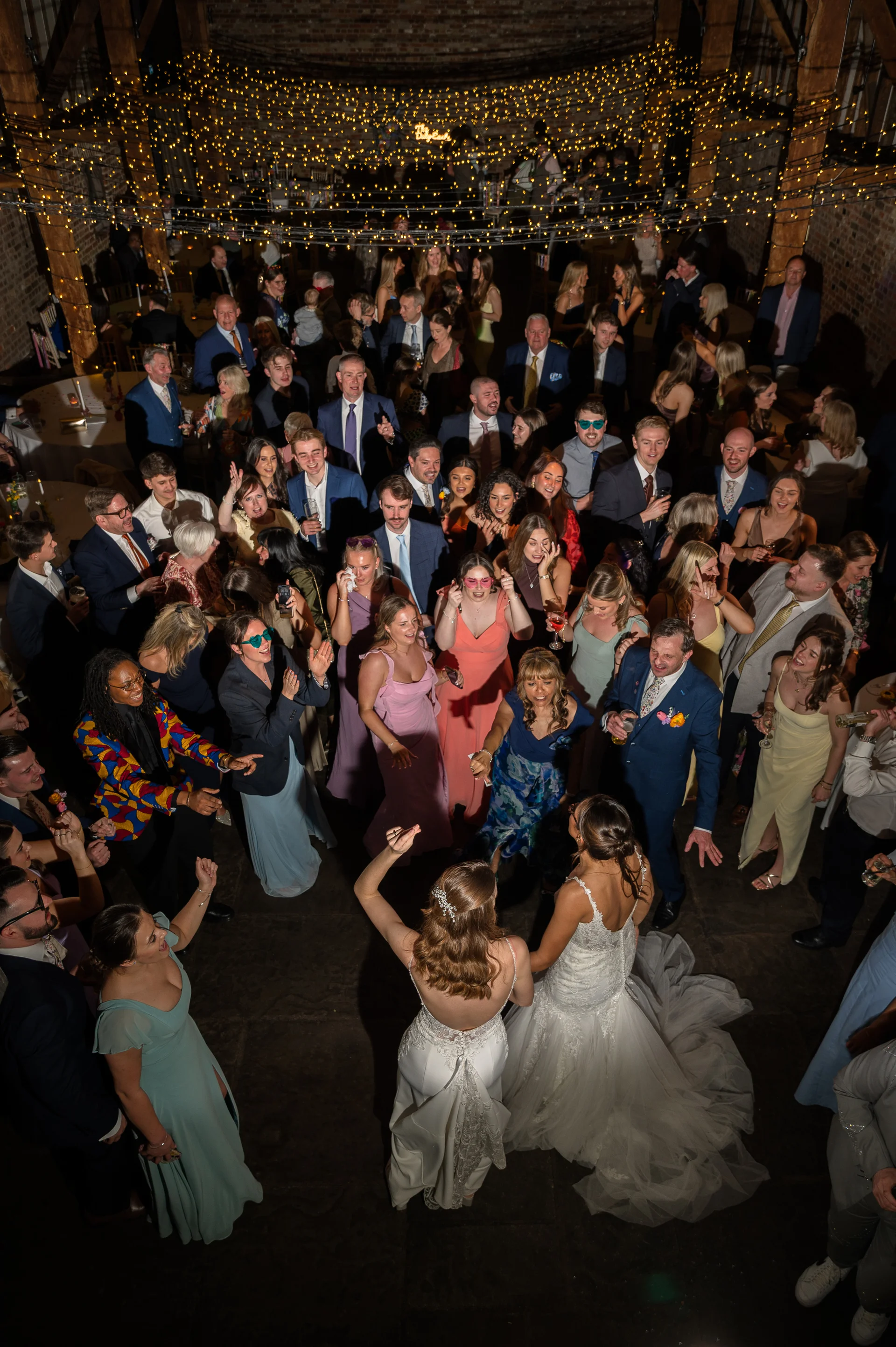 Two brides are captured from a high angle dancing surrounded by a large, joyful crowd of wedding guests inside a rustic barn illuminated by a canopy of fairy lights.