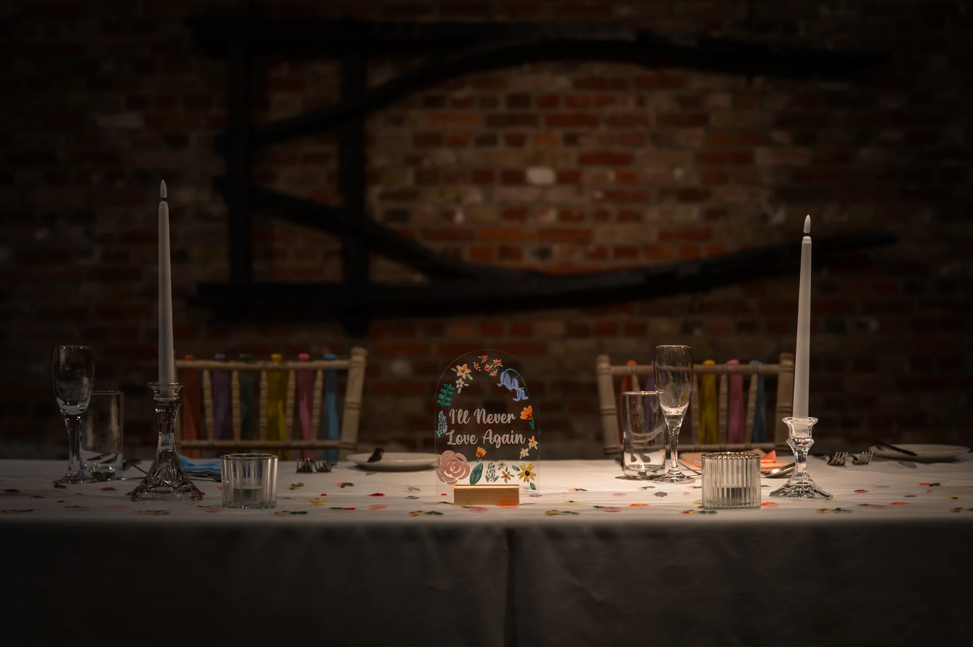 A moody, candlelit wedding reception table features an acrylic table number sign set against a rustic exposed brick wall.