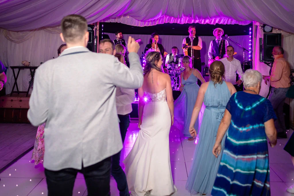 Newlyweds and wedding guests enjoy dancing on an illuminated dance floor inside a marquee featuring live band music and dramatic purple lighting.