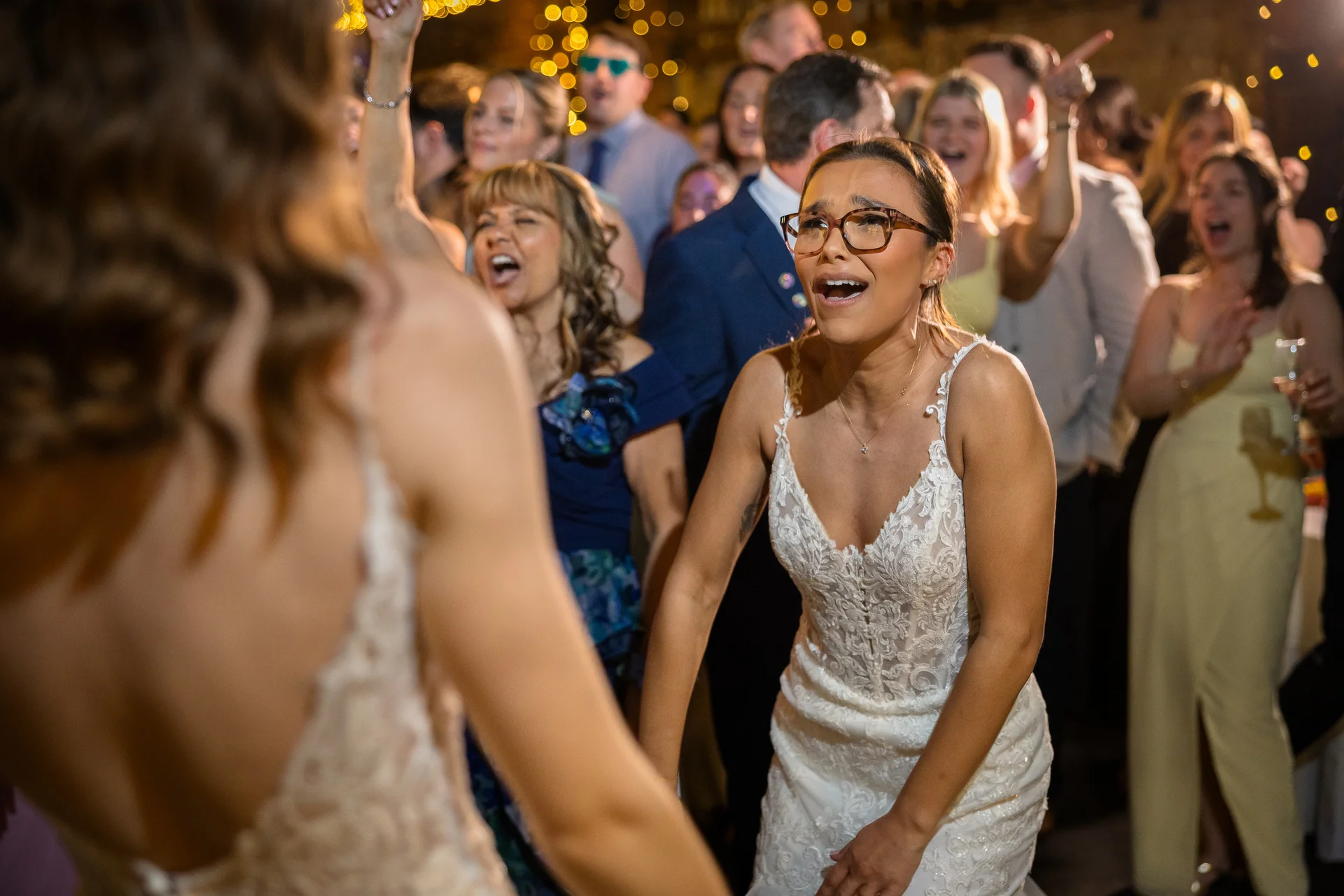 A joyful bride wearing glasses and a lace gown sings and dances enthusiastically on a packed dance floor surrounded by cheering wedding guests.