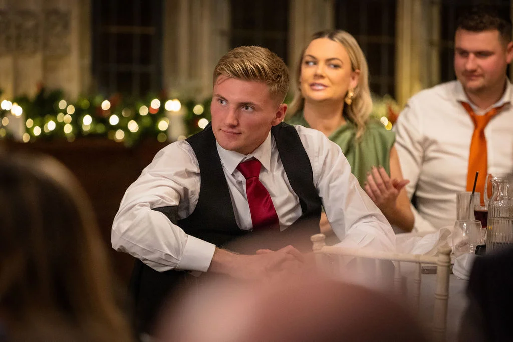 A young man wearing a waistcoat and red tie sits attentively at a wedding reception table while listening to speeches.