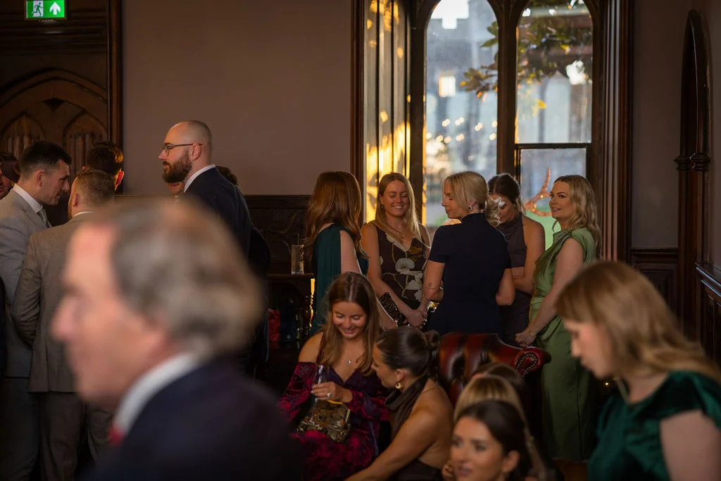 Wedding guests mingle during a drinks reception inside a historic country house with large gothic windows.