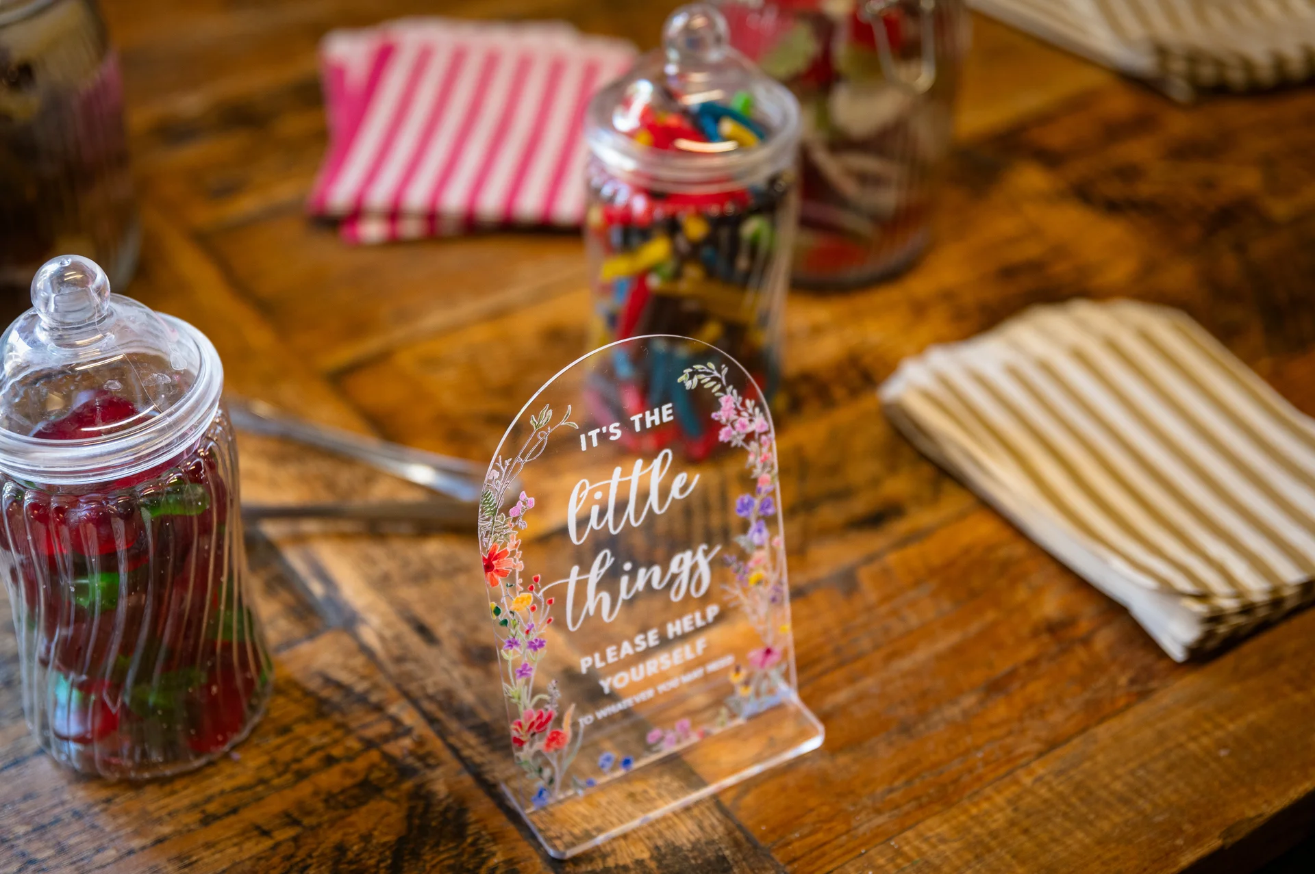 An acrylic sweet table sign featuring floral illustrations sits on a rustic wooden table alongside glass jars filled with retro pick and mix sweets.