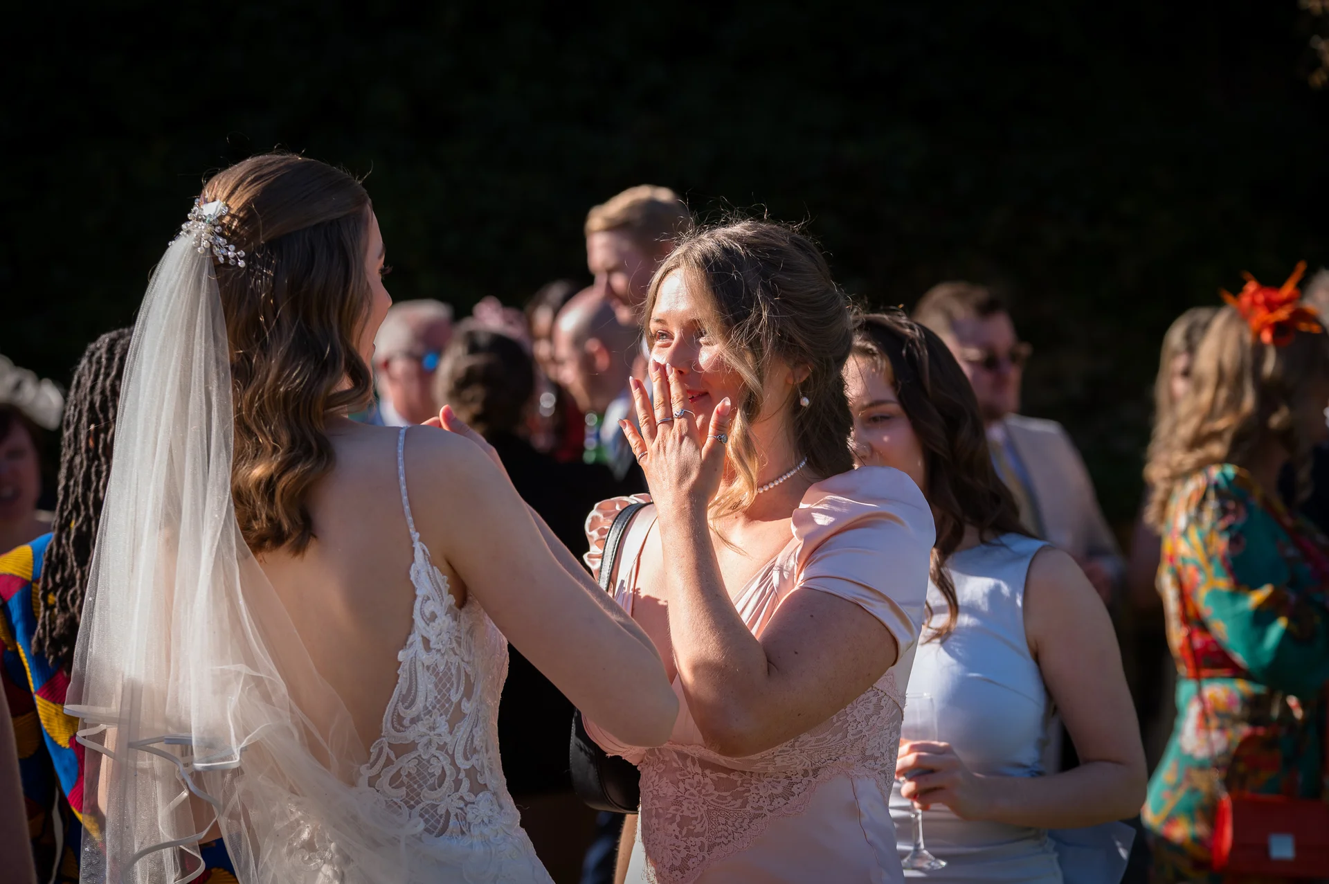 An emotional guest in a pink dress covers her mouth in surprise while interacting with a radiant bride wearing a lace gown and veil during an outdoor drinks reception.
