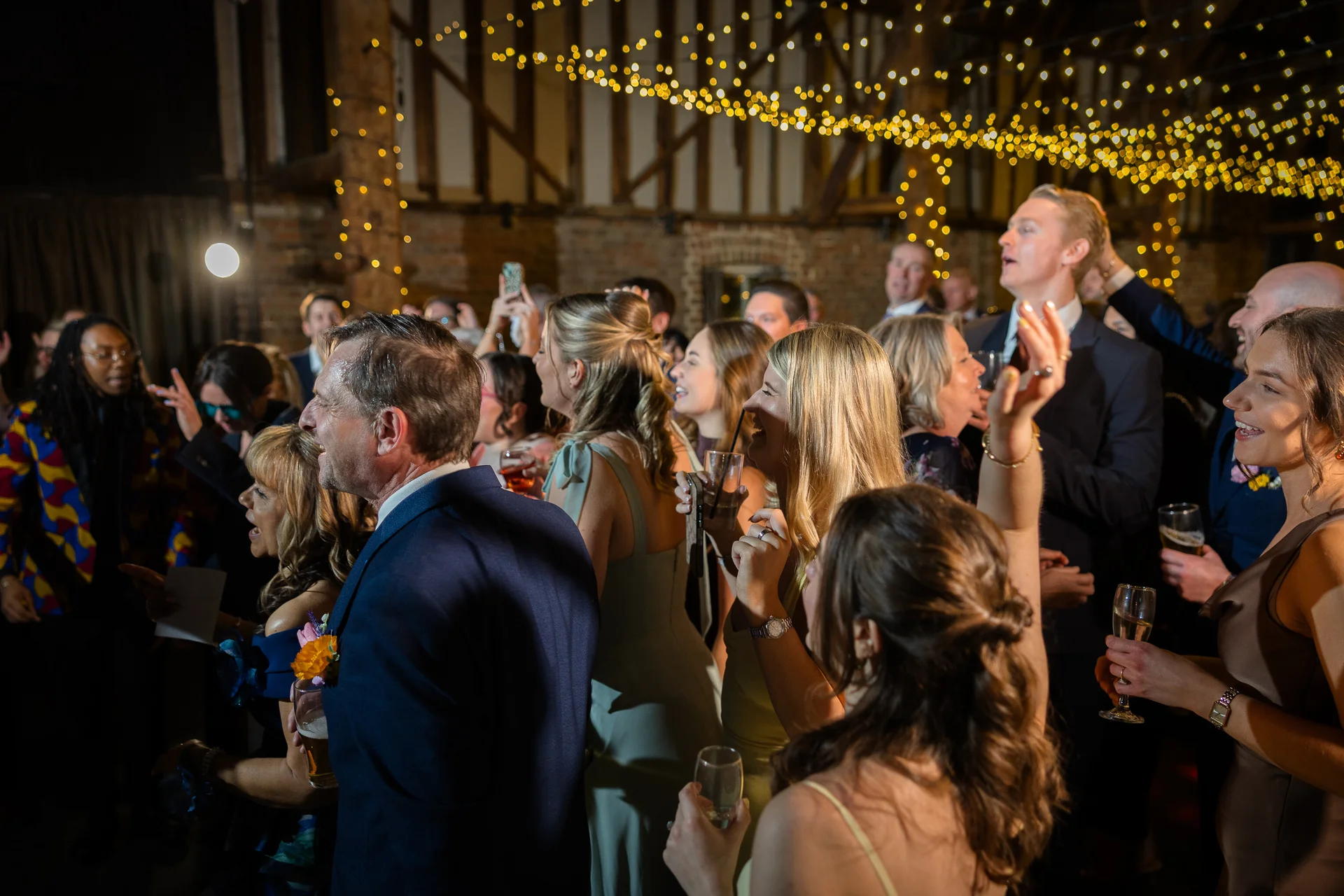 A joyful crowd of wedding guests celebrate and socialise under a canopy of fairy lights inside an oak-framed rustic barn venue.