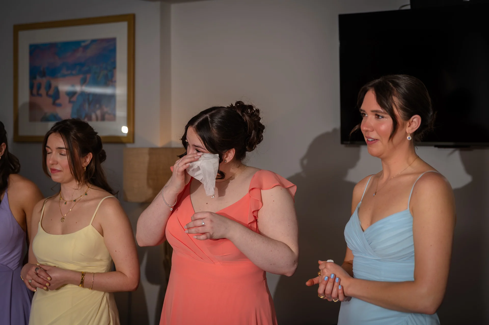 A bridesmaid in a coral pink dress wipes away tears of emotion with a tissue, flanked by fellow bridesmaids wearing mismatched pastel dresses.
