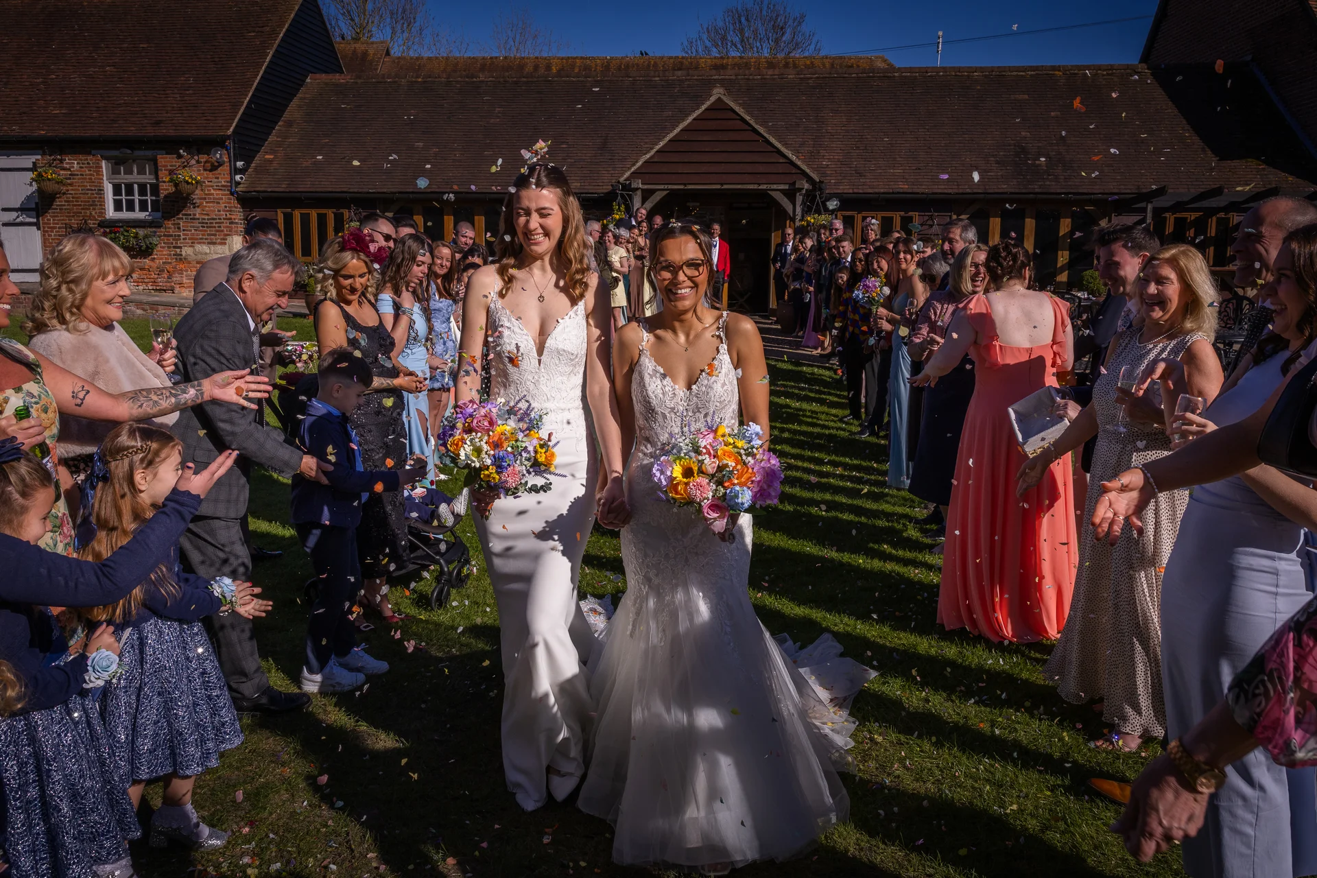 Two smiling brides walk hand in hand down an outdoor aisle during their sunny barn wedding while guests joyfully throw colorful confetti.