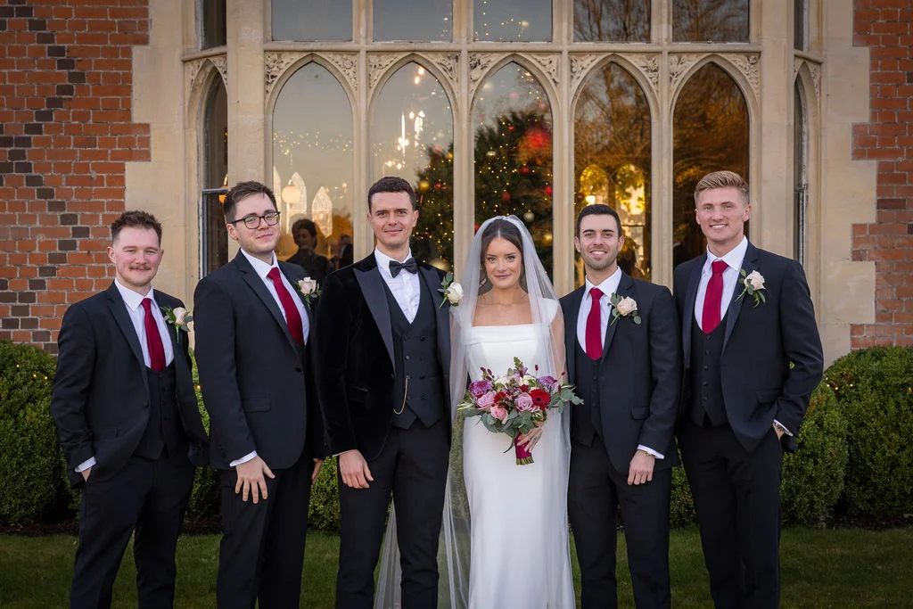 The bride and groom pose alongside four groomsmen outside a grand historic building featuring large stone-arched windows and red brickwork.