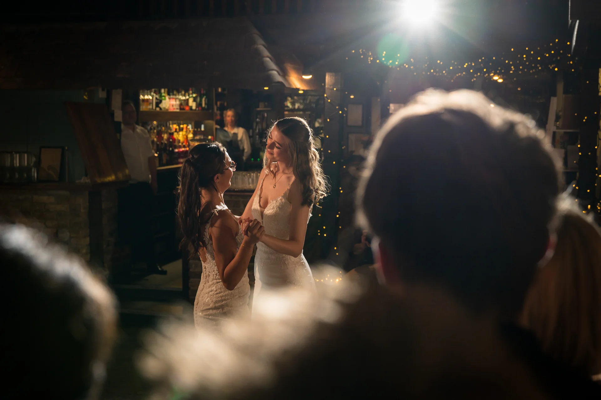 Two brides share a tender moment dancing together during their evening reception in a dimly lit pub illuminated by overhead string lights.