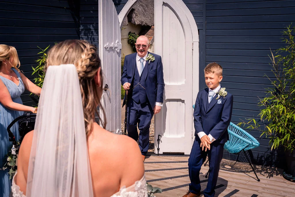 A bride in a strapless gown with a veil looks on as her father and a page boy in blue suits prepare for the ceremony outside white arched doors.