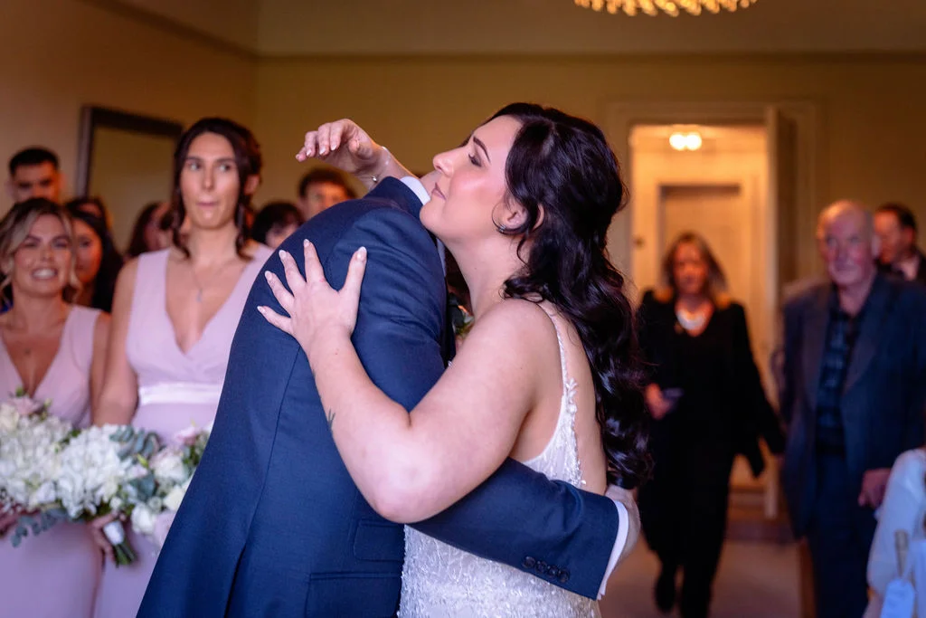 A newly married couple share an emotional embrace and kiss at the altar, surrounded by family and wedding party guests inside a hotel venue.