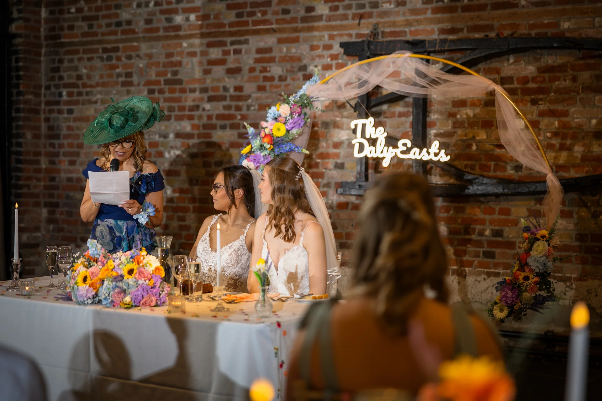 Two brides listen intently at the top table while a guest in a green fascinator delivers a heartfelt speech during the wedding reception.