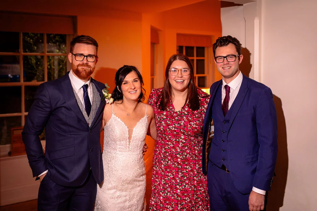 A happy wedding party poses indoors with the bride in a lace gown flanked by men in blue suits and a guest in a floral dress.