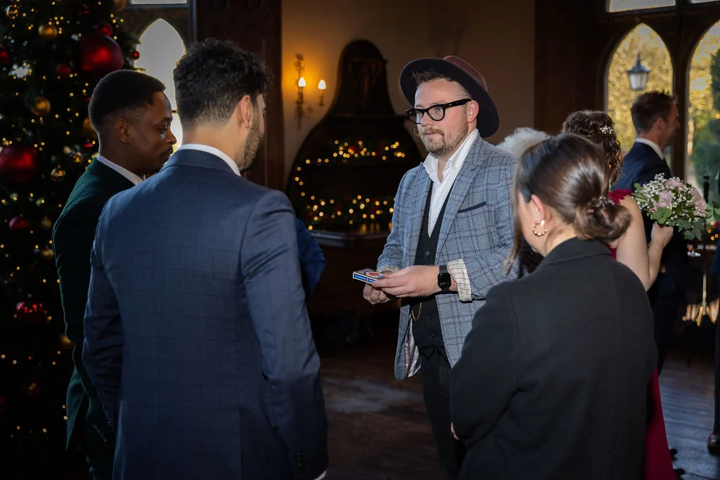 A close-up magician performs card tricks for a group of wedding guests gathered near a decorated Christmas tree during the reception.