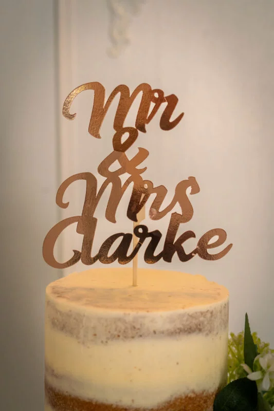 A close-up of a single-tier semi-naked wedding cake adorned with a custom rose gold Mr & Mrs Clarke cake topper.