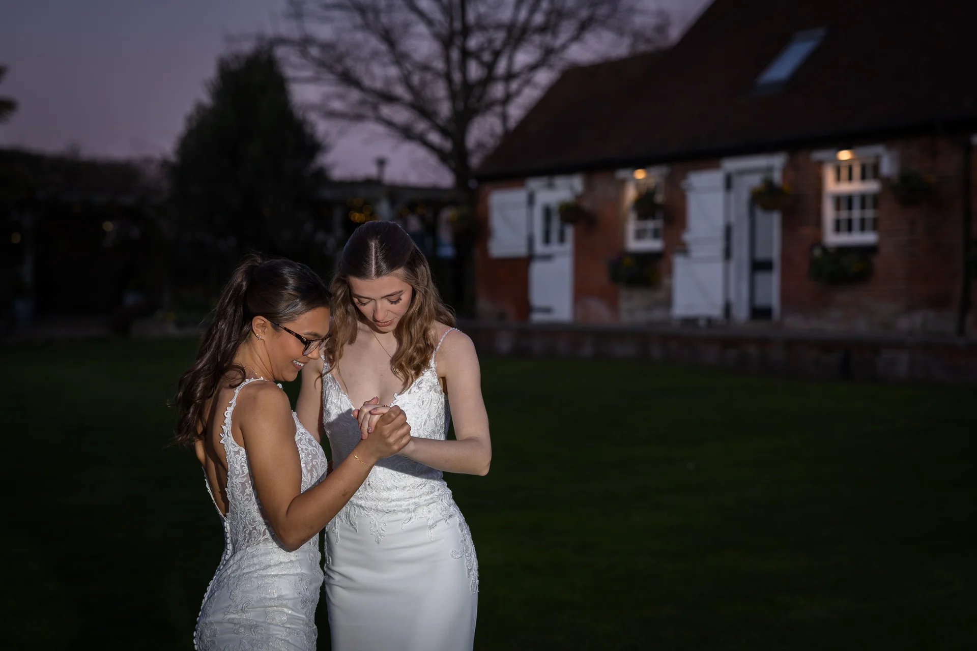 Two brides in white lace wedding dresses share a tender moment holding hands on the lawn outside a brick barn at twilight.