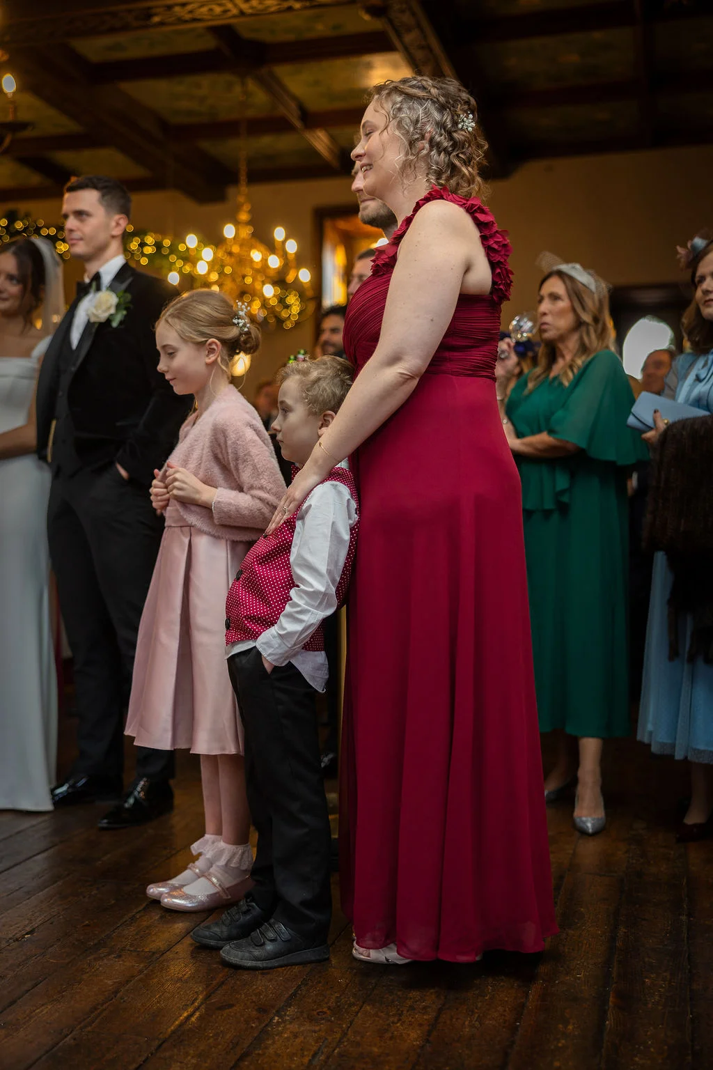 Wedding guests including a young flower girl and page boy stand watching the ceremony inside a rustic country house venue with wooden floorboards.