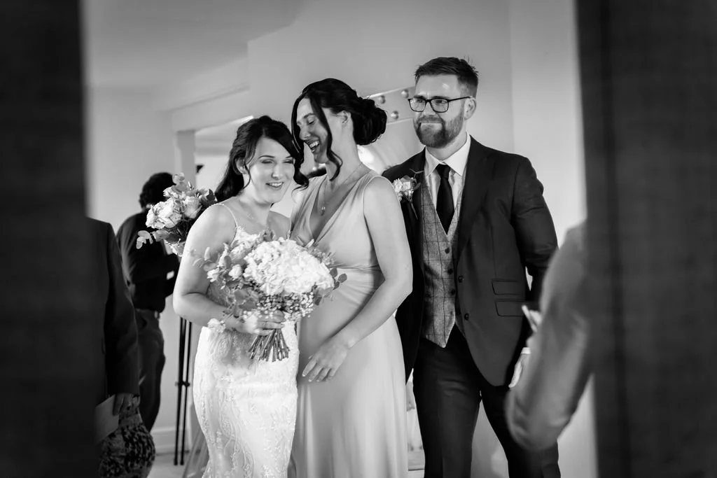 A smiling bride holding a bouquet stands alongside a bridesmaid and a groom in a tweed waistcoat during wedding preparations.