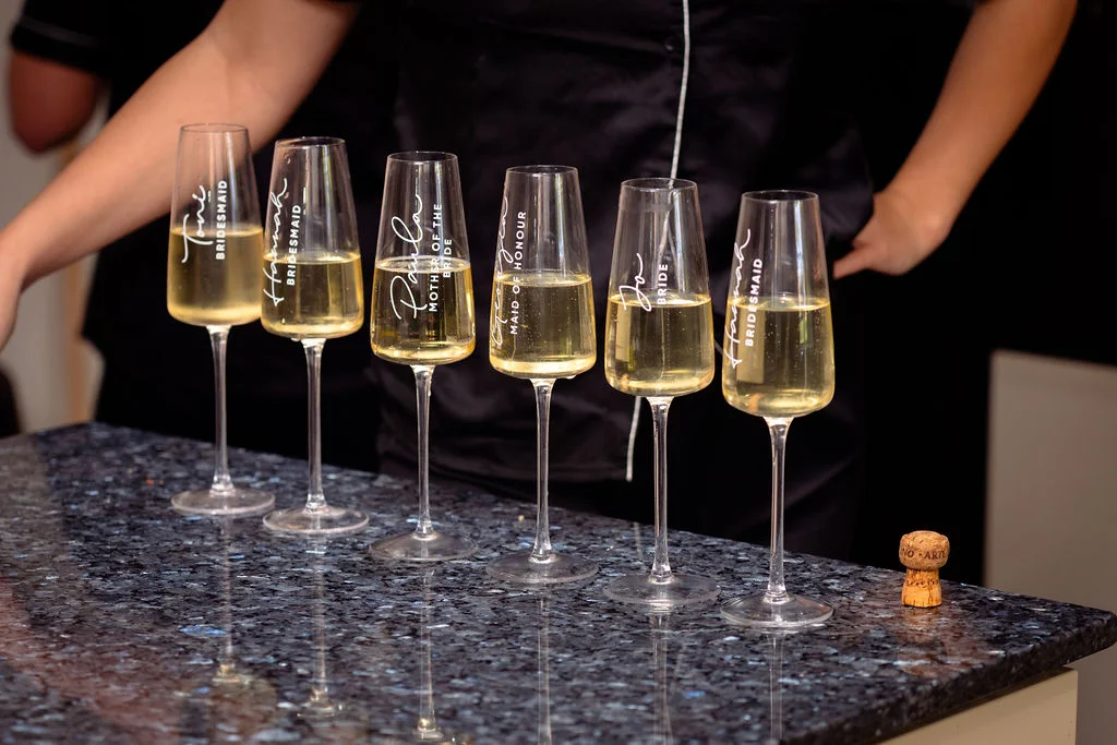 A line of personalised champagne flutes filled with sparkling wine sits on a dark marble countertop during bridal preparations.