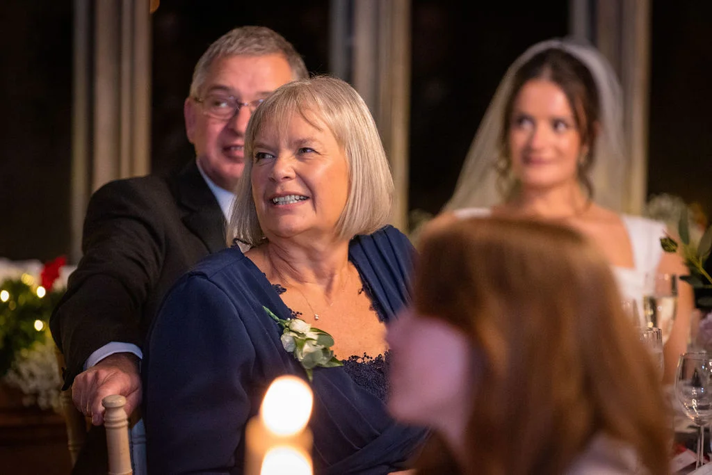 The mother of the bride smiles happily during wedding speeches while seated at the reception table alongside other guests and the bride.