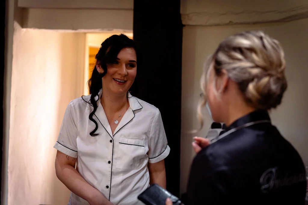 Two women laughing and chatting indoors while wearing matching satin pyjamas during morning bridal preparations.