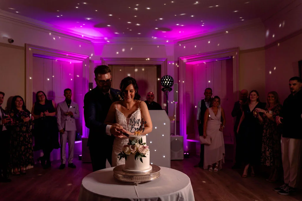 The newly married couple cut their two-tier wedding cake together surrounded by guests in a room illuminated with vibrant pink and white disco lighting.