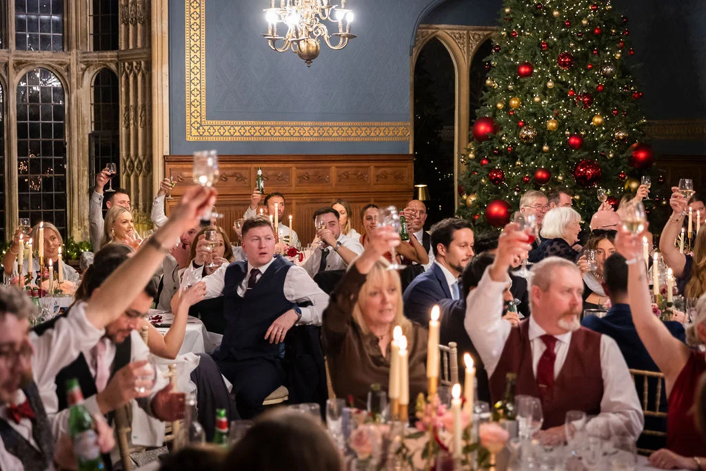 Wedding guests raise their glasses in a toast during the reception inside a stately reception room decorated with a large Christmas tree.