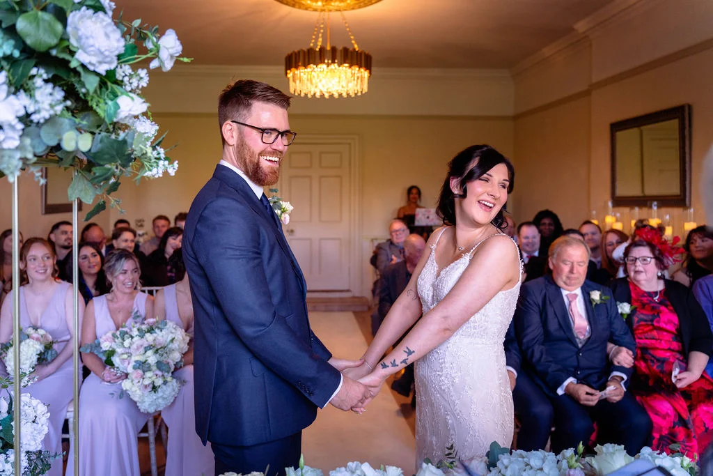 A smiling bride and groom hold hands during their indoor civil ceremony, surrounded by seated wedding guests and floral decorations.