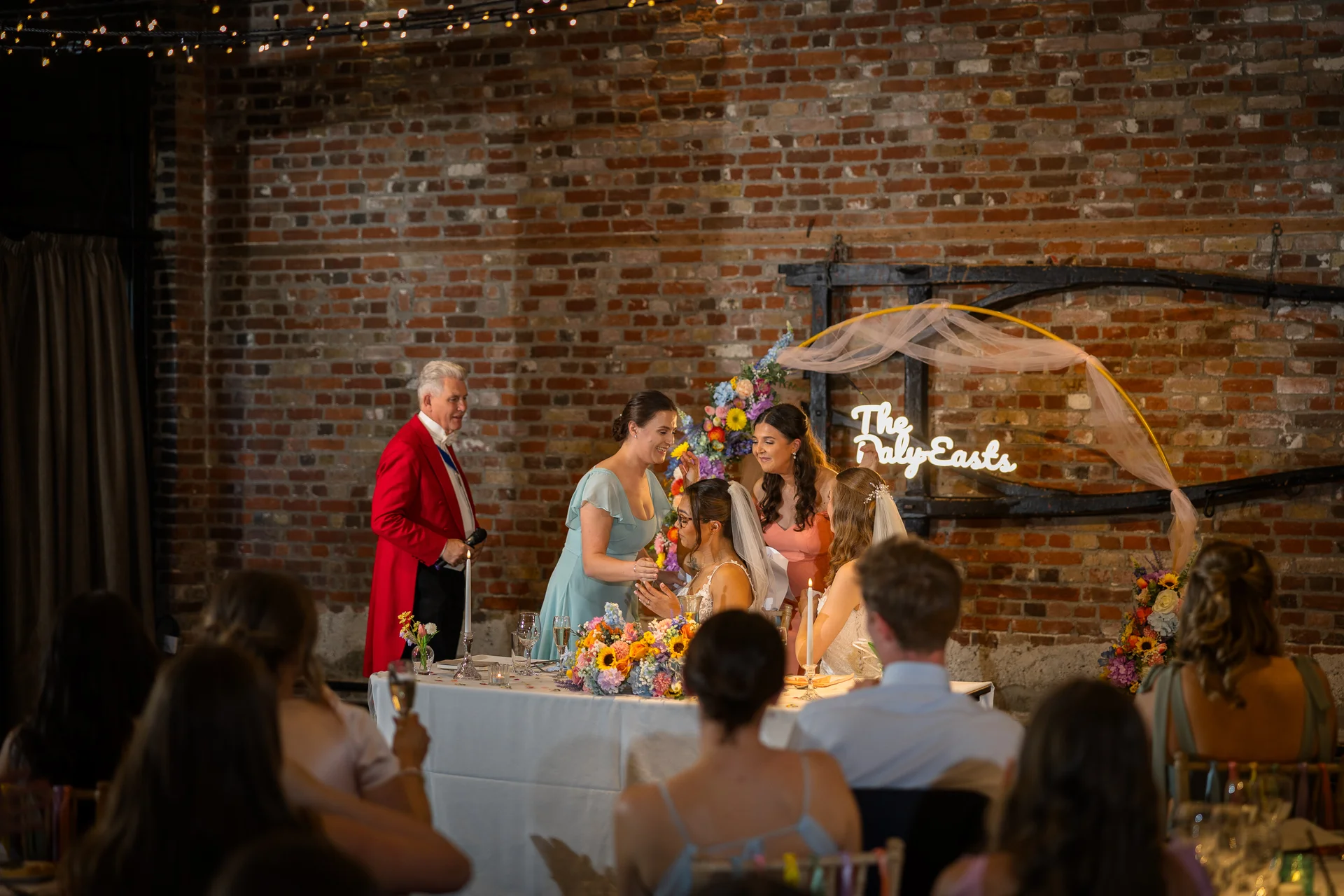 A toastmaster in a bright red tailcoat watches over the top table during wedding speeches inside an industrial venue with exposed brick walls and a glowing neon sign.