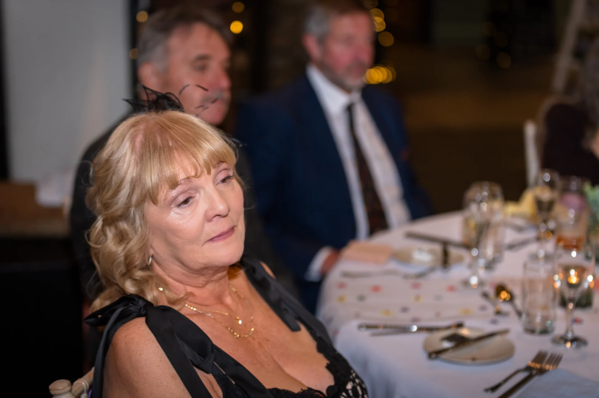 A mature female guest wearing a black evening dress sits at a beautifully set wedding breakfast table while listening to speeches.