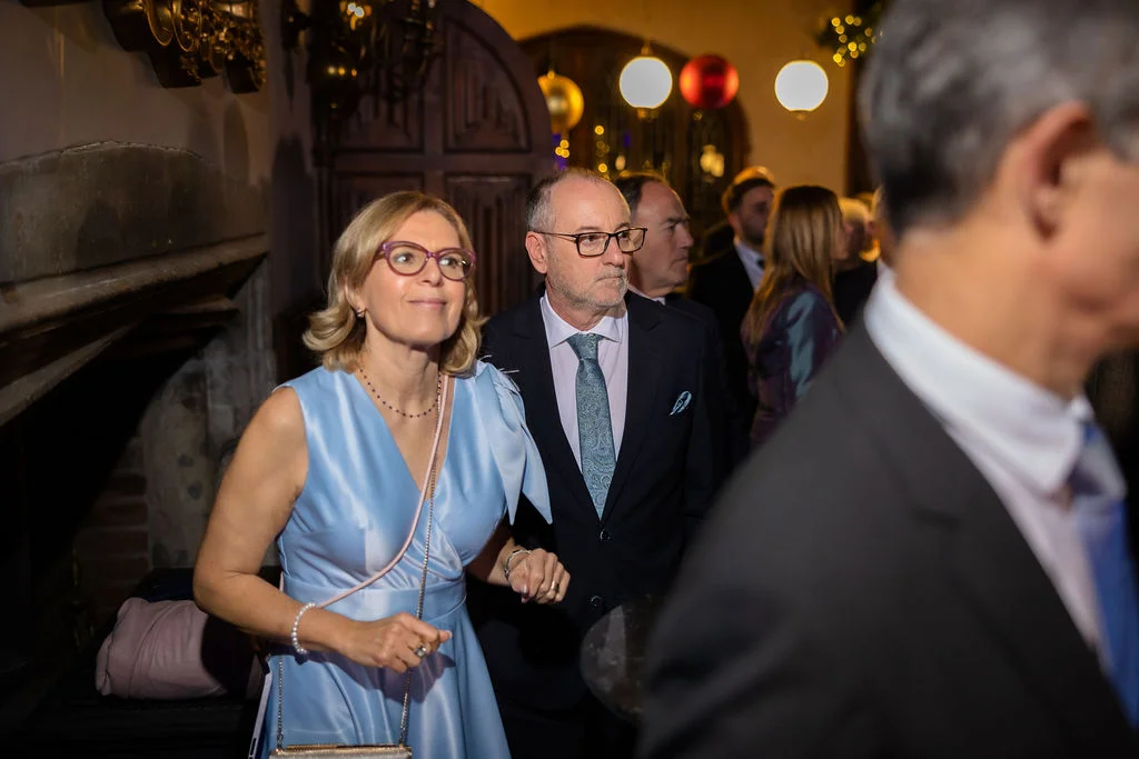 An elegantly dressed couple mingles with guests inside a venue during a wedding reception, with the woman wearing a light blue dress and glasses.