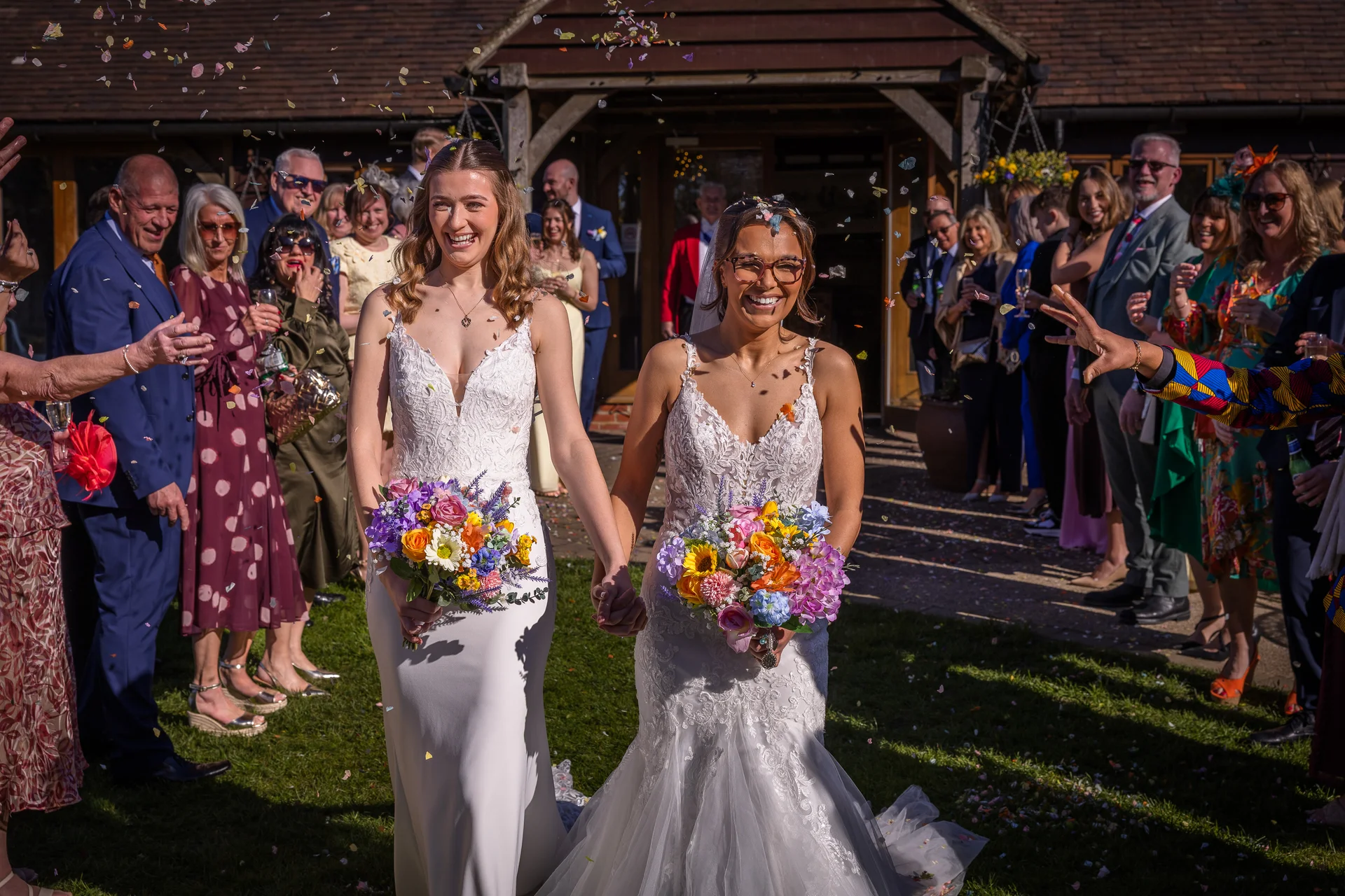 Two smiling brides walk hand in hand through an outdoor confetti shower as wedding guests cheer and celebrate around them in the sunshine.